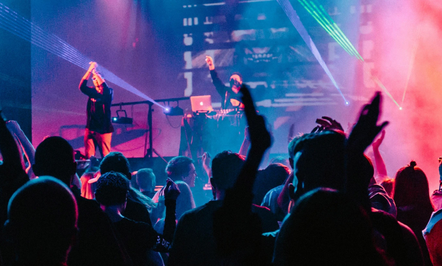 A crowd with their hands up at a gig, under pink and blue stage lights